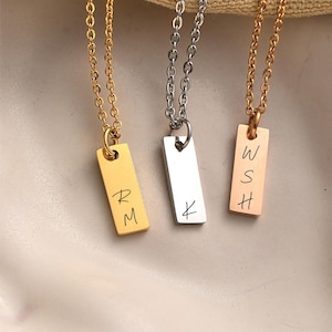 May include: Three gold, silver, and rose gold necklaces with personalized rectangular pendants. The gold pendant has the initials "RM" engraved on it, the silver pendant has the letter "K" engraved on it, and the rose gold pendant has the initials "WSH" engraved on it.