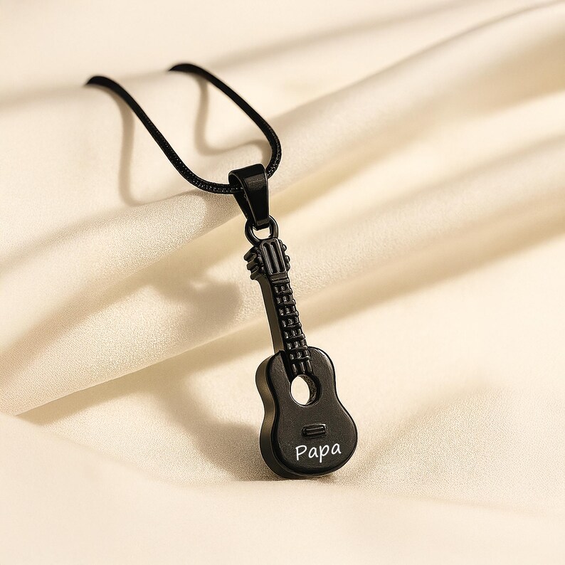 K&ouml;nnte beinhalten: Eine schwarze gitarrenf&ouml;rmige Anh&auml;ngerkette mit einer d&uuml;nnen schwarzen Kordel. Der Gitarrenk&ouml;rper ist mit dem Wort Papa in wei&szlig;er Schrift graviert. Der Anh&auml;nger hat einen detaillierten Hals, eine Kopfplatte und ein rundes Schallloch in Mattschwarz.