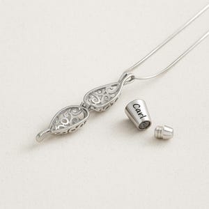 Pu&ograve; includere: Una collana con ciondolo in metallo color argento caratterizzata da un design a goccia traforato con intricati motivi a volute. Accanto al ciondolo si trova un piccolo tappo cilindrico in metallo inciso con il nome Carl, dotato di vite.