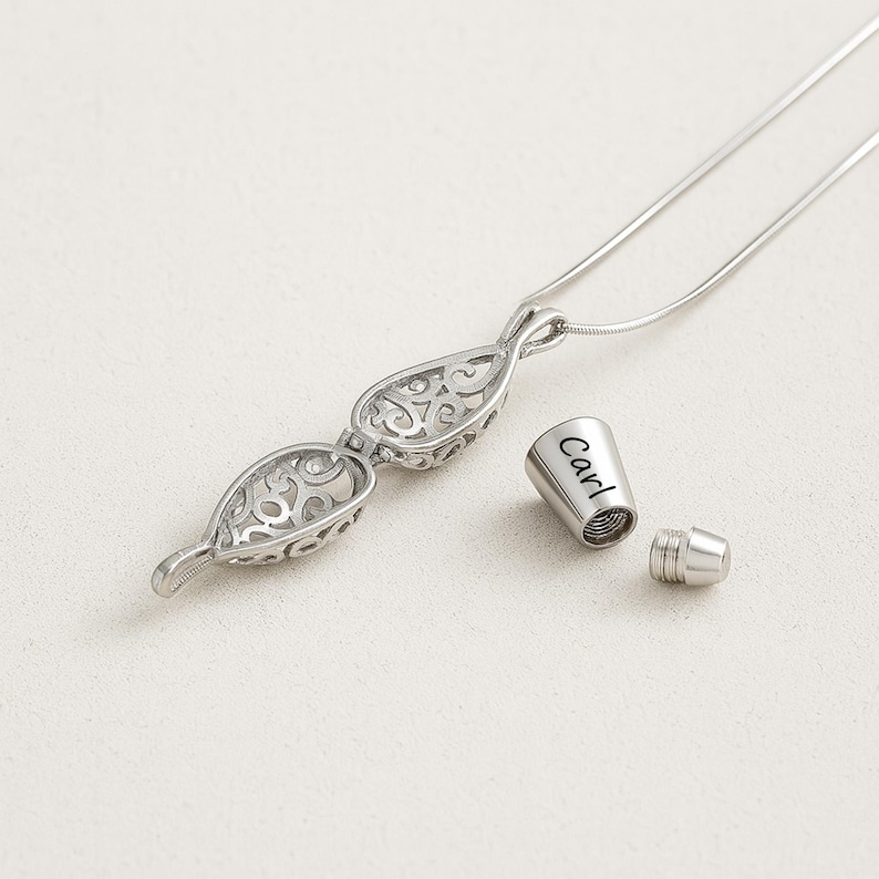 Pu&ograve; includere: Una collana con ciondolo in metallo color argento caratterizzata da un design a goccia traforato con intricati motivi a volute. Accanto al ciondolo si trova un piccolo tappo cilindrico in metallo inciso con il nome Carl, dotato di vite.
