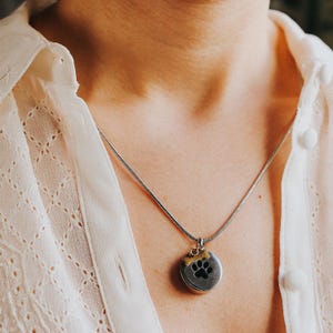 May include: A silver necklace with a round pendant featuring a black paw print and a small gold-colored bone. The pendant hangs from a delicate silver chain. The necklace is worn over a white blouse.