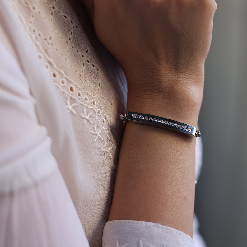 A silver stainless steel bracelet with a Cuban link chain and a curved bar adorned with sparkling crystals. The elegant and sophisticated design makes it a perfect accessory for both casual and formal wear, adding a touch of brilliance to any outfit.