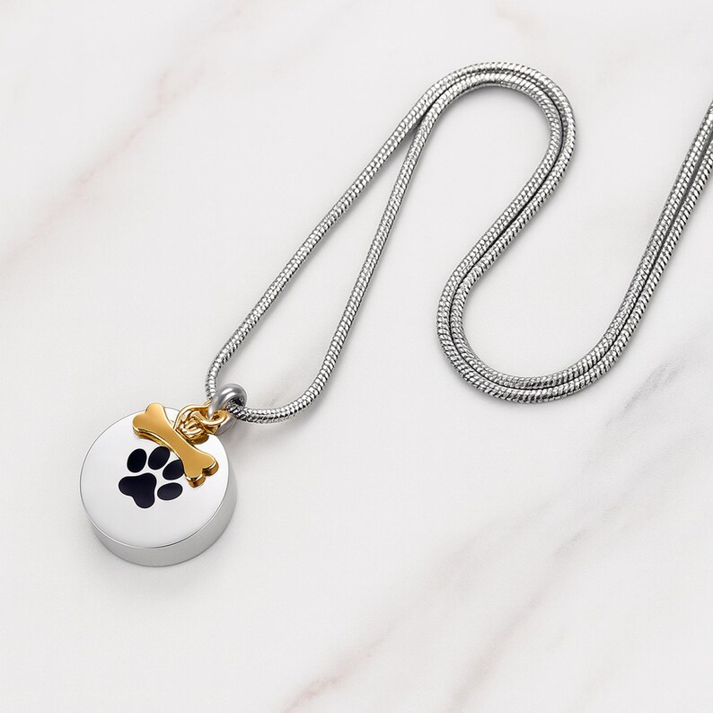 May include: A silver-colored pet memorial necklace with a round pendant featuring a black paw print. A gold-colored bone charm hangs above the pendant. The necklace has a silver chain.
