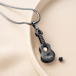 K&ouml;nnte beinhalten: Ein gitarrenf&ouml;rmiger Anh&auml;nger aus schwarzem Metall mit einem hohlen Korpus und einem detaillierten Hals. Das Wort "Papa" ist auf die Vorderseite des Gitarrenkorpus graviert. Ein kleines, separates schwarzes Gewindeschraubenteil liegt daneben.