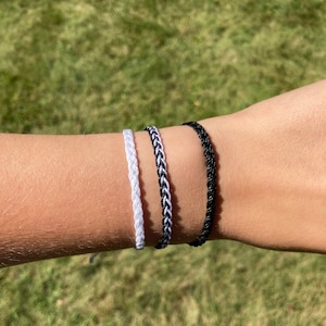 May include: Three braided friendship bracelets in black, white, and black and white. The bracelets are worn on a person's wrist.