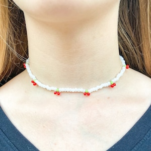 Peut inclure: Un collier ras de cou en perles blanches avec des perles rouges et vertes formant un motif de cerise.