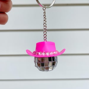 May include: A pink cowboy hat with rhinestones sits atop a silver disco ball keychain.