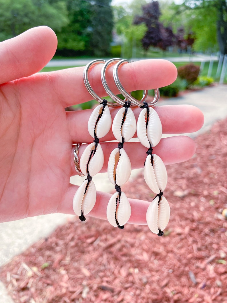 May include: A keychain with three strands of white seashells strung on black cord. Each strand has four seashells and is attached to a silver key ring.