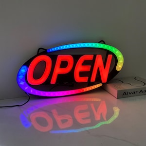 以下が含まれることがあります： 黒い楕円形のフレームの付いた、光る「OPEN」サイン。 「OPEN」の文字は明るい赤色で、カラフルなLEDライトのボーダーで囲まれています。 サインの反射が下の表面に見えます。 右側には本があります。