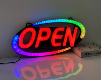 May include: Illuminated "OPEN" sign with a black oval frame. The word "OPEN" is in bright red, surrounded by a border of colorful LED lights. The sign's reflection is visible on the surface below. A book is on the right side.