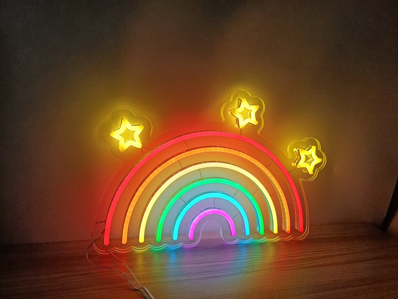 May include: A rainbow neon sign with five glowing yellow stars. The rainbow is made of six different colors: red, orange, yellow, green, blue, and purple. The sign is on a wooden surface.
