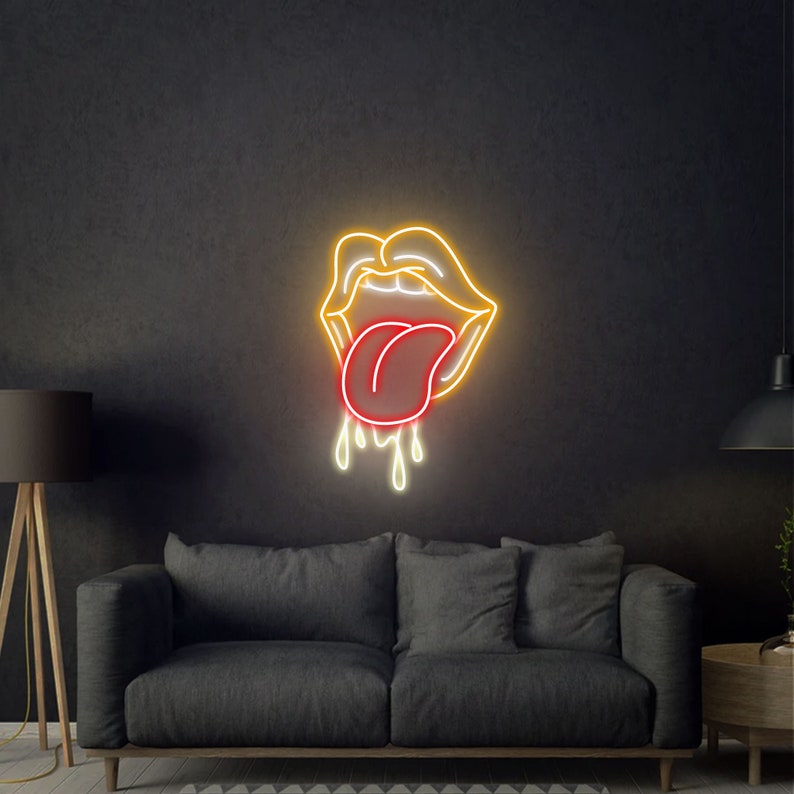 May include: Neon sign depicting a pair of lips with a red tongue sticking out and dripping. The lips are outlined in yellow, with the tongue and drips in red and white. The sign is mounted on a dark gray wall above a gray sofa.
