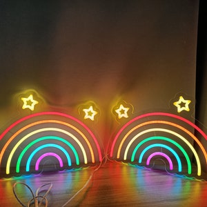 May include: Two rainbow neon signs with stars on a wooden surface. Each sign features a multi-colored rainbow with a yellow, orange, red, green, blue, indigo, and violet color scheme.