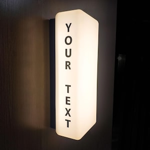Puede incluir: Una lámpara de pared rectangular e iluminada con las palabras "YOUR TEXT" impresas verticalmente en negro. La lámpara tiene una apariencia suave y brillante y está montada en una pared oscura.