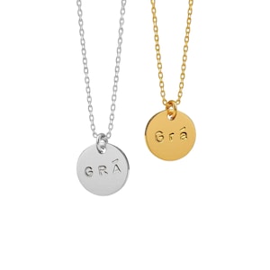 Könnte beinhalten: Zwei Halsketten mit dünnen Ketten und runden Anhängern. Ein Anhänger ist silberfarben und mit dem Text "GRÁ" in Großbuchstaben geprägt. Der andere Anhänger ist goldfarben und mit dem Text "Grá" in gemischter Schreibweise auf der runden Fläche geprägt.