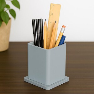 Könnte beinhalten: Ein grauer Schreibtisch-Organizer gefüllt mit schwarzen Stiften, Bleistiften, einem blauen Marker und einem Holzlineal. Der Organizer ist quadratisch mit einer kleinen Basis und steht auf einer dunkelbraunen Oberfläche. Eine Topfpflanze ist im Hintergrund.