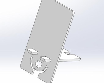 May include: A grey phone stand with a smiling face cutout design. The stand is made from a single piece of material and features a slot for holding a phone upright. The stand has a rectangular base for stability.