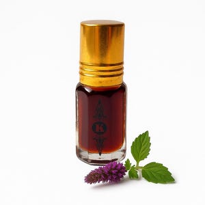 Puede incluir: Una pequeña botella de vidrio transparente con tapa dorada, llena de líquido ámbar oscuro. La botella tiene un emblema negro con la letra "K" en la parte delantera. Una ramita de hojas verdes y una flor morada están junto a la botella.