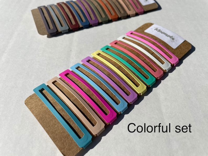 May include: A set of 10 colourful hair clips in various pastel shades, including pink, blue, yellow, and green. The clips are arranged on a brown cardboard backing with the word "Kisamede" printed on it. The text "Colorful set" is printed below the clips.