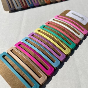 May include: A set of 12 colourful hair clips in pastel shades of blue, pink, yellow, green, and white. The clips are arranged in a row on a brown cardboard backing. The word "Kismet" is printed on the backing.