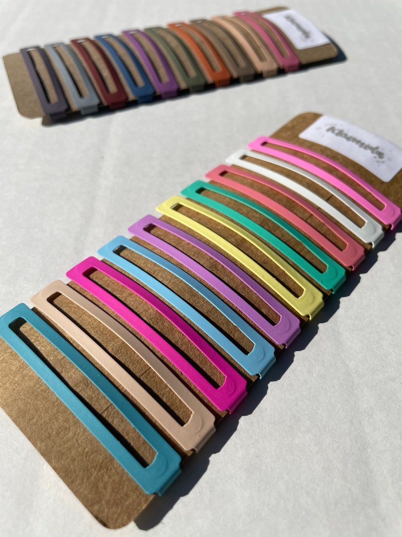 May include: A set of 12 colourful hair clips in pastel shades of blue, pink, yellow, green, and white. The clips are arranged in a row on a brown cardboard backing. The word "Kismet" is printed on the backing.