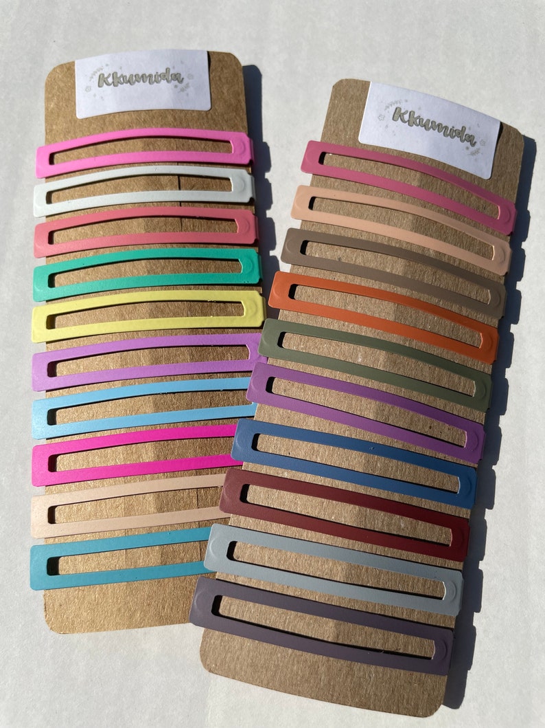 May include: Two sets of colourful hair clips on a brown cardboard backing. The clips are arranged in rows and are made of metal with a variety of colours, including pink, blue, green, yellow, and purple. The backing has the word "Khairvida" printed on it.