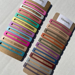 May include: Two sets of colourful hair clips on a brown cardboard backing. The clips are arranged in rows, with each row featuring a different colour. The top set features pastel colours, while the bottom set features more muted tones. The packaging includes the word "Klunvida" printed on each card.