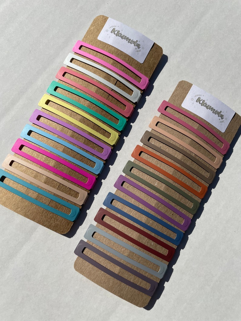 May include: Two sets of colourful hair clips on a brown cardboard backing. The clips are arranged in rows, with each row featuring a different colour. The top set features pastel colours, while the bottom set features more muted tones. The packaging includes the word "Klunvida" printed on each card.