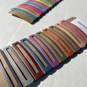 May include: Two sets of hair clips, one set of 12 pastel coloured clips and one set of 10 muted coloured clips. The clips are arranged in rows on a brown cardboard backing.