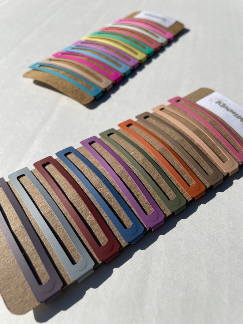 May include: Two sets of hair clips, one set of 12 pastel coloured clips and one set of 10 muted coloured clips. The clips are arranged in rows on a brown cardboard backing.
