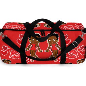 May include: Red duffel bag featuring a repeating pattern of white hearts, lips, and two cartoon figures in red dresses. The bag has black handles and a shoulder strap. The bag is designed for travel or gym use.