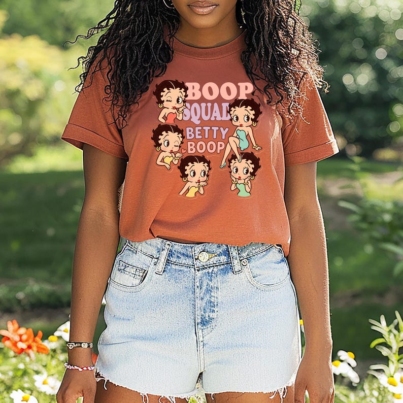 May include: A rust-colored t-shirt featuring a graphic of Betty Boop and the text "Boop Squad Betty Boop." The design includes multiple images of Betty Boop in various poses. The model is wearing denim shorts.