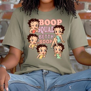 May include: Olive green t-shirt featuring a graphic of Betty Boop in various poses, with the text "Boop Squad Betty Boop" in pink and orange. The design includes multiple Betty Boop illustrations in different outfits.
