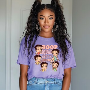May include: A lavender t-shirt featuring a graphic of Betty Boop and the text "Boop Squad Betty Boop". The design includes multiple Betty Boop illustrations in various poses. The shirt is paired with blue jeans.