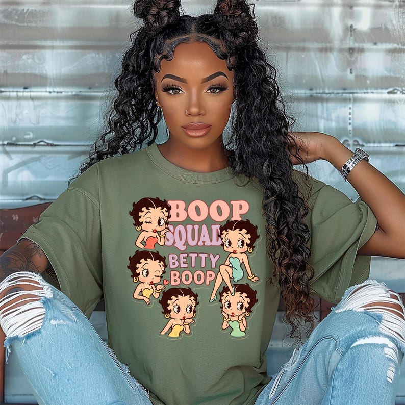 May include: Olive green t-shirt featuring a graphic of Betty Boop and the text "Boop Squad Betty Boop". The graphic includes multiple Betty Boop illustrations in various poses. The shirt is paired with light blue ripped jeans.