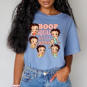 May include: A light blue t-shirt featuring a graphic of Betty Boop in various poses, with the text "Boop Squad Betty Boop" in a pink and orange font. The shirt is made of a soft material and has short sleeves.