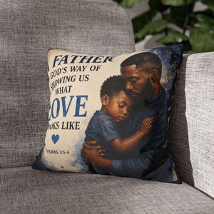 Op de afbeelding: Decoratief kussen met een afbeelding van een vader en kind in een omhelzing. De tekst "FATHER GOD'S WAY OF SHOWING US WHAT LOVE LOOKS LIKE" is blauw, met een hart. De achtergrond is crème met een blauw aquareleffect.