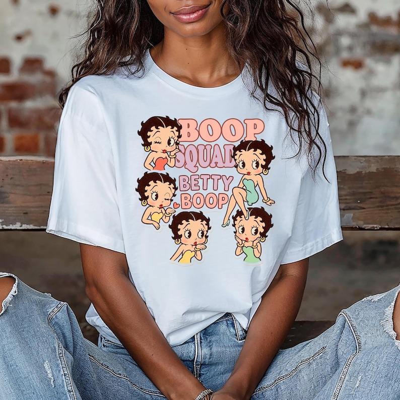 May include: White t-shirt featuring a graphic of Betty Boop and the text "Boop Squad Betty Boop" in pink and peach. The design includes multiple images of Betty Boop in various poses, wearing different colored outfits. The shirt has short sleeves.