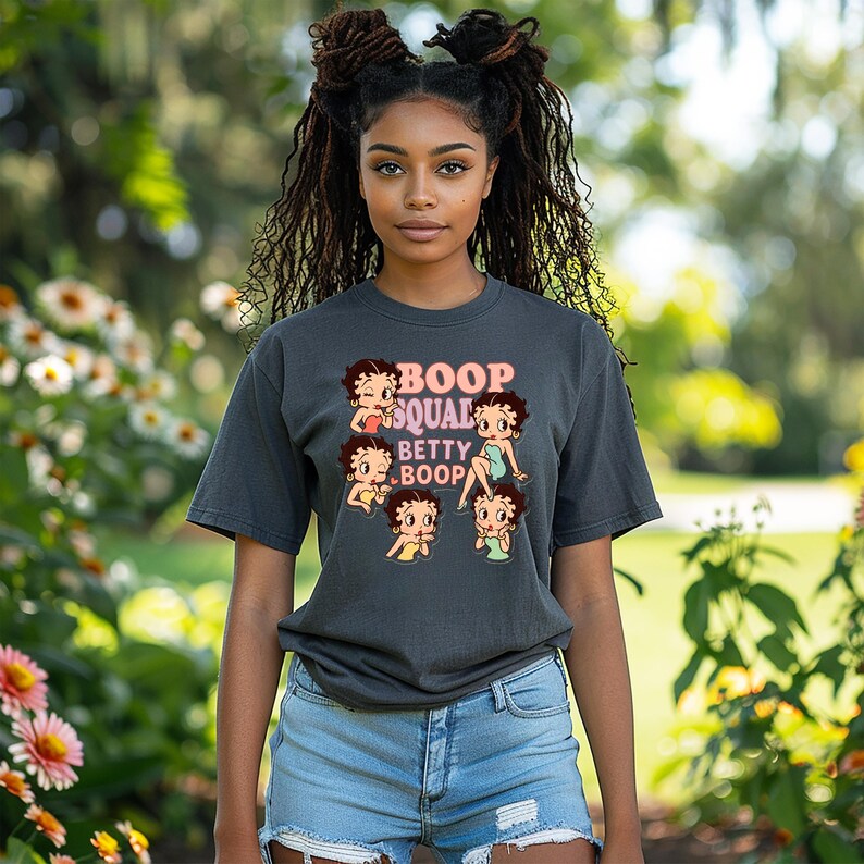 May include: A dark gray t-shirt featuring the text "Boop Squad Betty Boop" with multiple Betty Boop cartoon images. The shirt is paired with blue denim shorts. The background is a blurred outdoor scene with greenery and flowers.