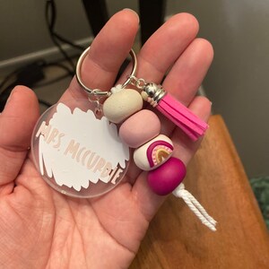 May include: A clear acrylic keychain with the text "Mrs. McCurdie" in a light brown color. The keychain has a beaded accent with pink, white, and magenta beads, a rainbow design, and a pink tassel.