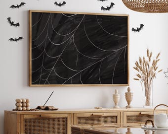 Op de afbeelding: Een zwart ingelijst canvas met een wit spinnenwebontwerp hangt aan een muur met zwarte vleermuisuitsparingen. De muur is wit en het canvas is ingelijst in zwart hout. Het canvas hangt boven een houten kast met een geweven rotan bovenkant.