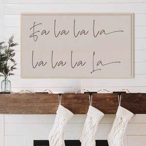 Puede incluir: Un letrero de madera blanco con las palabras "fa la la la la la la la" escritas en cursiva negra. El letrero está colgado sobre una chimenea con calcetines blancos colgados del manto.