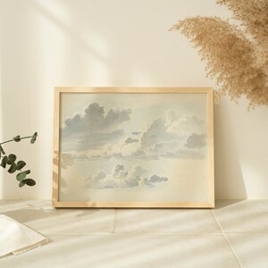 May include: A framed watercolor painting of a cloudy sky in shades of blue and gray. The painting is in a light wood frame and is hanging on a white wall.