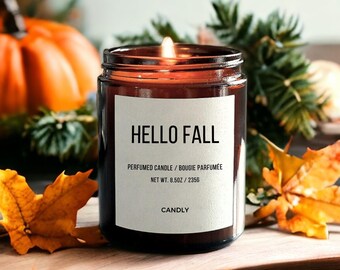 May include: A lit, scented candle in a dark glass jar with a white label that reads "HELLO FALL". The candle is surrounded by fall foliage, including orange leaves and a pumpkin. The candle weighs 8.5oz.