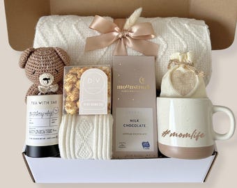Puede incluir: Una caja de regalo que contiene una manta de punto trenzado color crema con una cinta beige, un oso de ganchillo marrón, un recipiente de té, una caja de palomitas de caramelo, una barra de chocolate con leche Moonstruck y una taza crema y tostada #momlife.