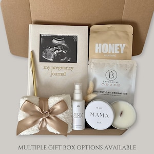May include: A gift box with a pregnancy theme, containing a journal with an ultrasound image, a pen, a small towel with a bow, a bottle of Mom Glow serum, a honey packet, a bath soak, and a candle. The box is beige.