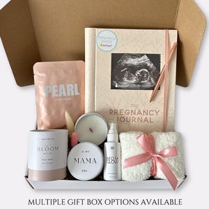 May include: A gift box containing a pregnancy journal, skincare products, a candle, and a plush towel tied with a pink ribbon. The journal features a sonogram image and the words "My Pregnancy Journal". The box also includes a bag of "Pearl Brightening" powder.
