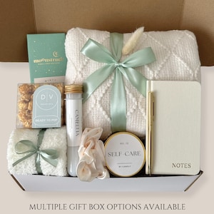May include: A gift box filled with self-care items. Includes a white blanket tied with a mint green ribbon, a notebook, a pen, a candle, a tube labeled "Camellia", popcorn, a scrunchie, and a small book. The box is labeled "Multiple Gift Box Options Available."