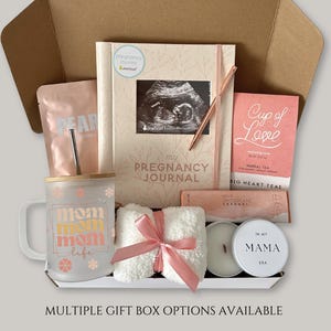 May include: A gift box containing a pregnancy journal with an ultrasound photo, a frosted glass mug with a bamboo lid and straw, a pink pouch, a box of herbal tea, a chocolate bar, a white fuzzy item with a pink ribbon, and a candle in a tin.