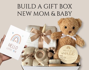 May include: A gift box filled with items for a new mom and baby. The box contains a white blanket, a teddy bear, a wooden rattle, a wooden ring, a brush, a card that says "Hello Little One" with a rainbow design, and a wooden sign that says "Hello World" with space to write the baby's name, date and time of birth, weight, and length.